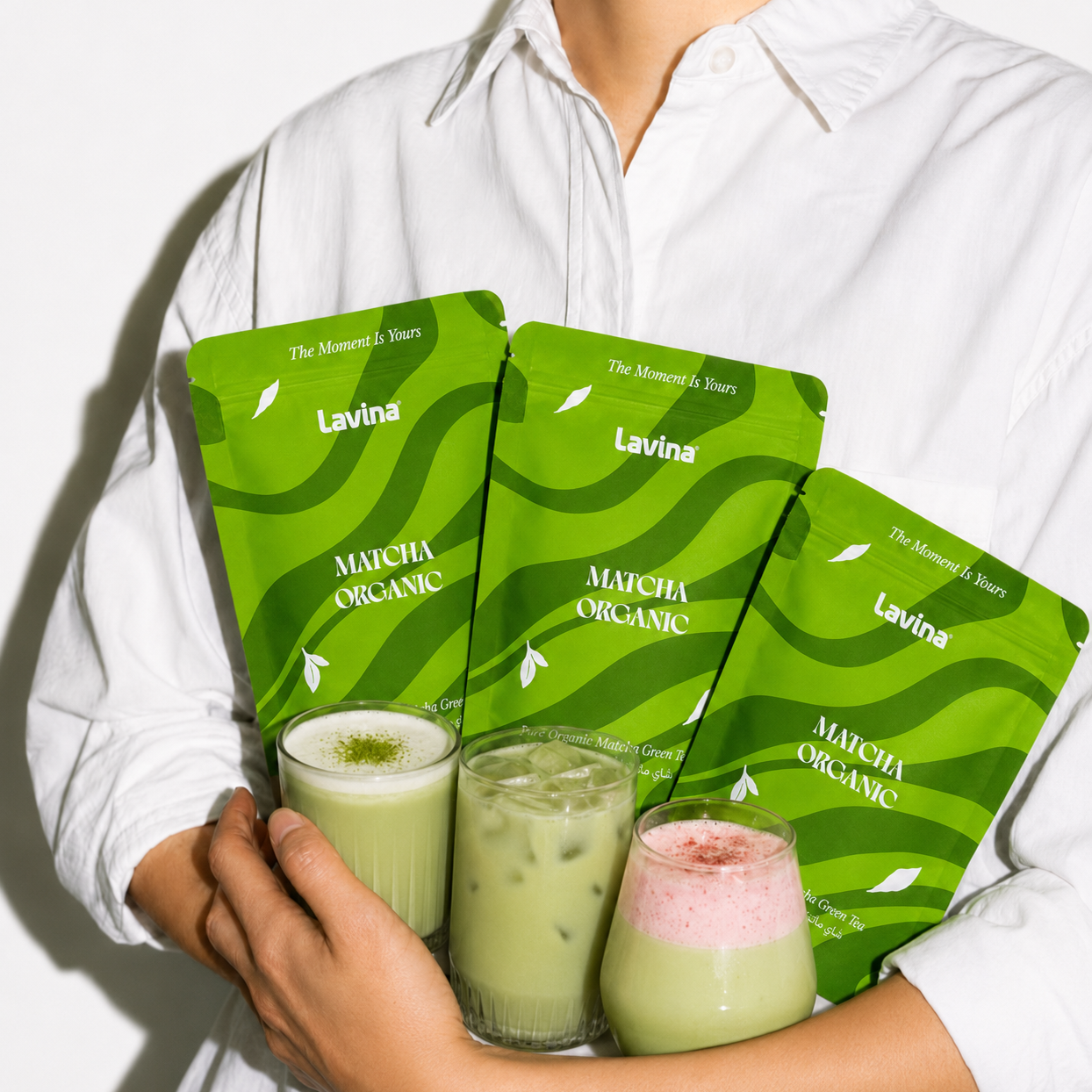 A person in a white button-up shirt holds three green Lavina Organic matcha pouches and three prepared matcha drinks in glasses against a clean white background.